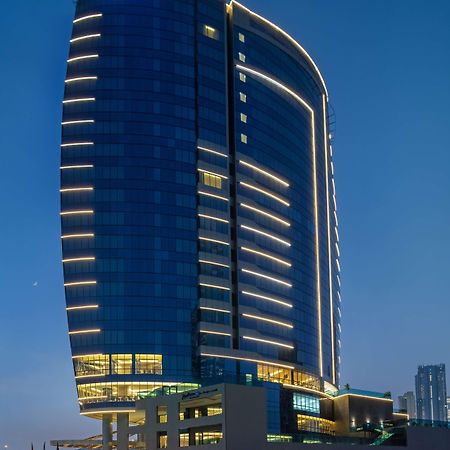 Radisson Blu Hotel, Dubai Canal View Exterior photo The image shows a modern skyscraper with a sleek, curved design. The building is primarily covered in glass, reflecting the evening sky, and features illuminated lines along its surface, enhancing its architectural appeal. The lighting creates a stri