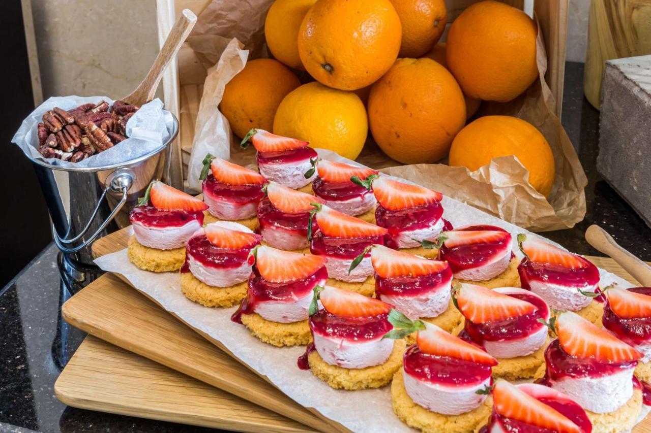 Radisson Blu Hotel, Dubai Canal View Exterior photo The photo displays a delightful spread of desserts. In the foreground, there are small dessert bites arranged on a wooden board. Each bite features a golden crust topped with a layer of cream, a strawberry slice, and a drizzle of red fruit sauce. Beh