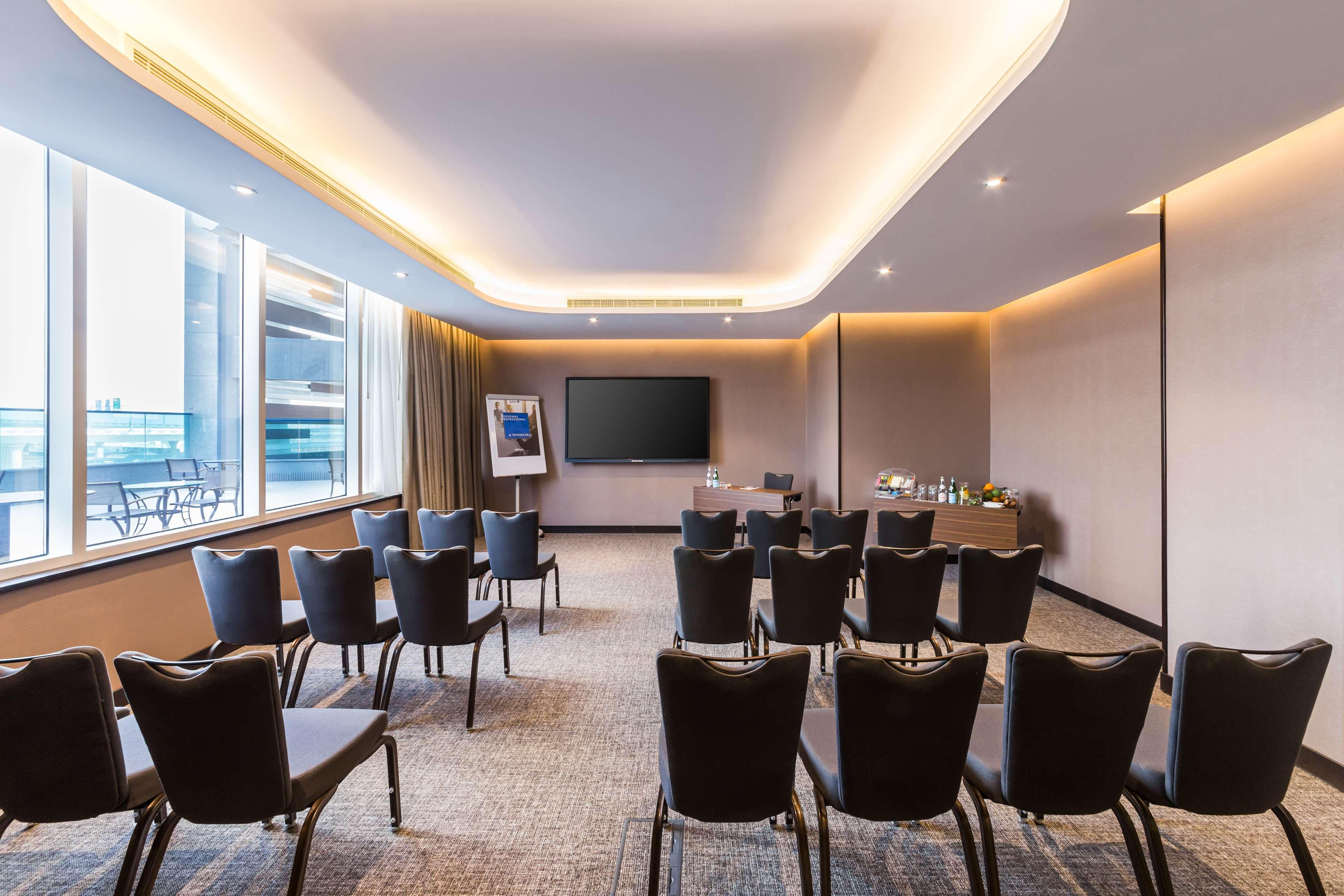 Radisson Blu Hotel, Dubai Canal View Exterior photo The photo shows a modern conference room designed for meetings or presentations. It features several rows of black chairs arranged facing a large wall-mounted screen. The room has windows that allow natural light to enter, and there are subtle ambien