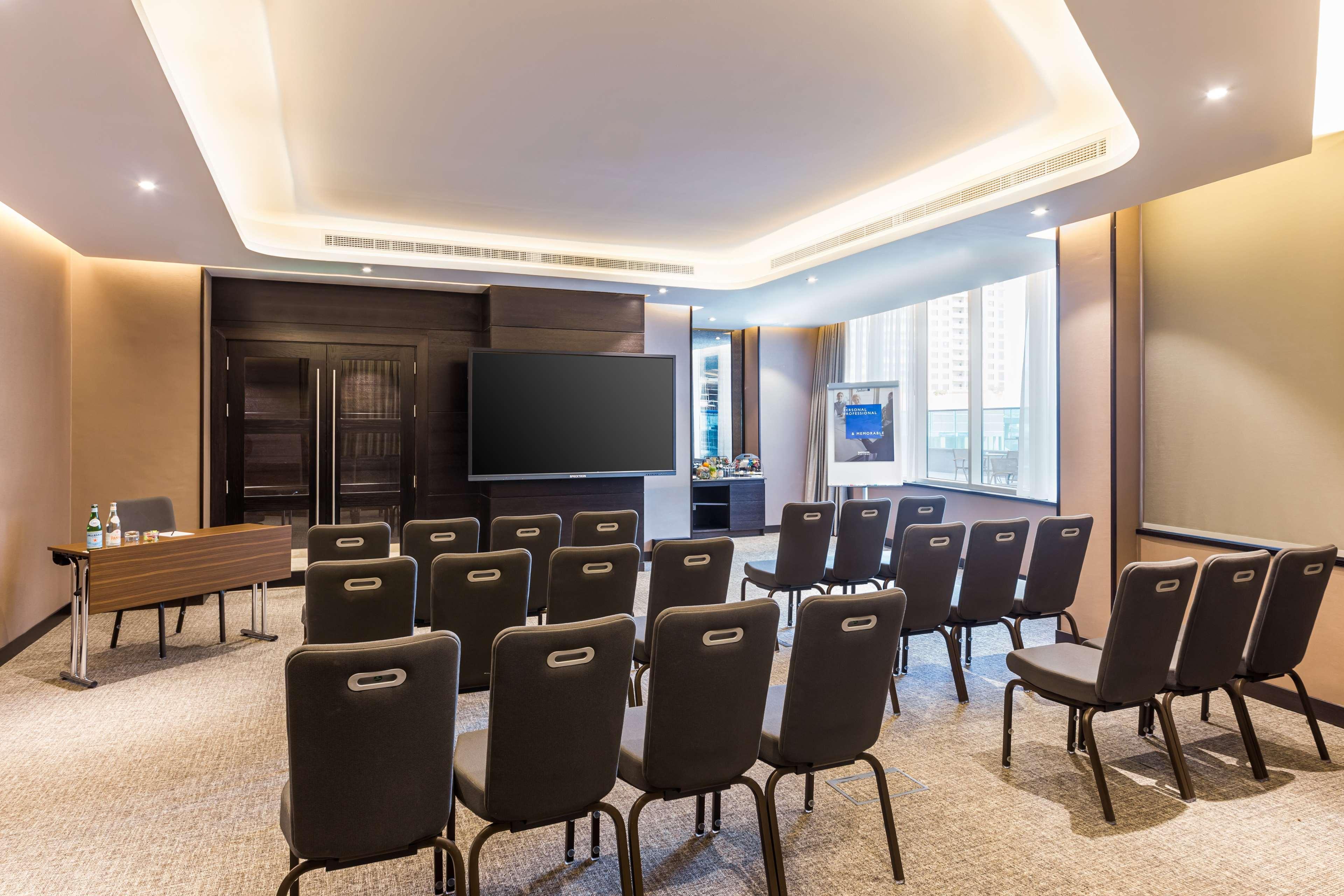 Radisson Blu Hotel, Dubai Canal View Exterior photo The photo shows a modern conference room set up for a meeting or presentation. There are several rows of black chairs arranged facing a large screen or monitor mounted on the wall. The room is well-lit with natural light coming through the windows. T