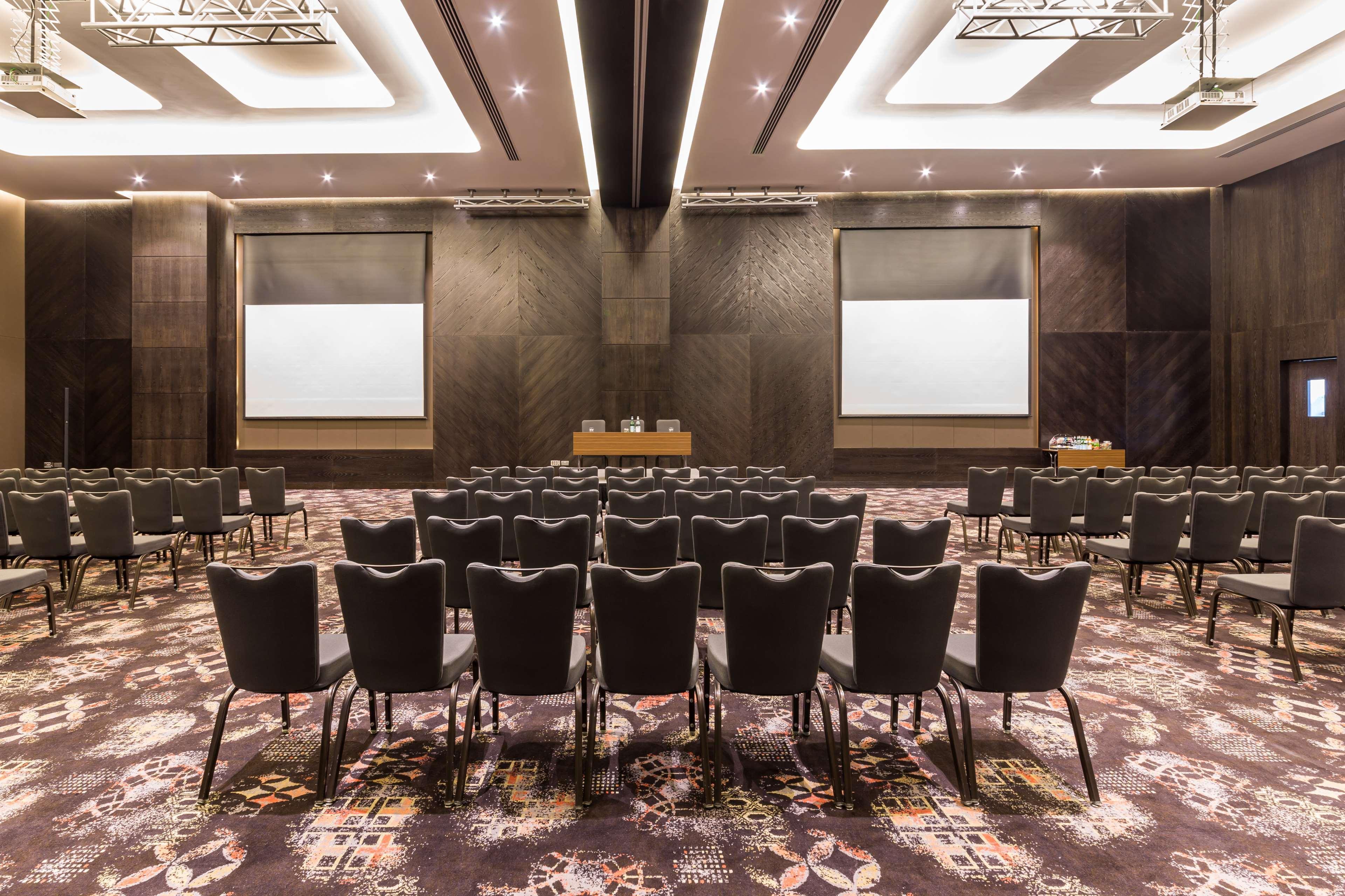 Radisson Blu Hotel, Dubai Canal View Exterior photo The image shows a conference room or meeting space set up for an event. There are numerous rows of black chairs arranged facing a stage or presentation area. In the front, there are two large screens on the walls, likely intended for displaying prese