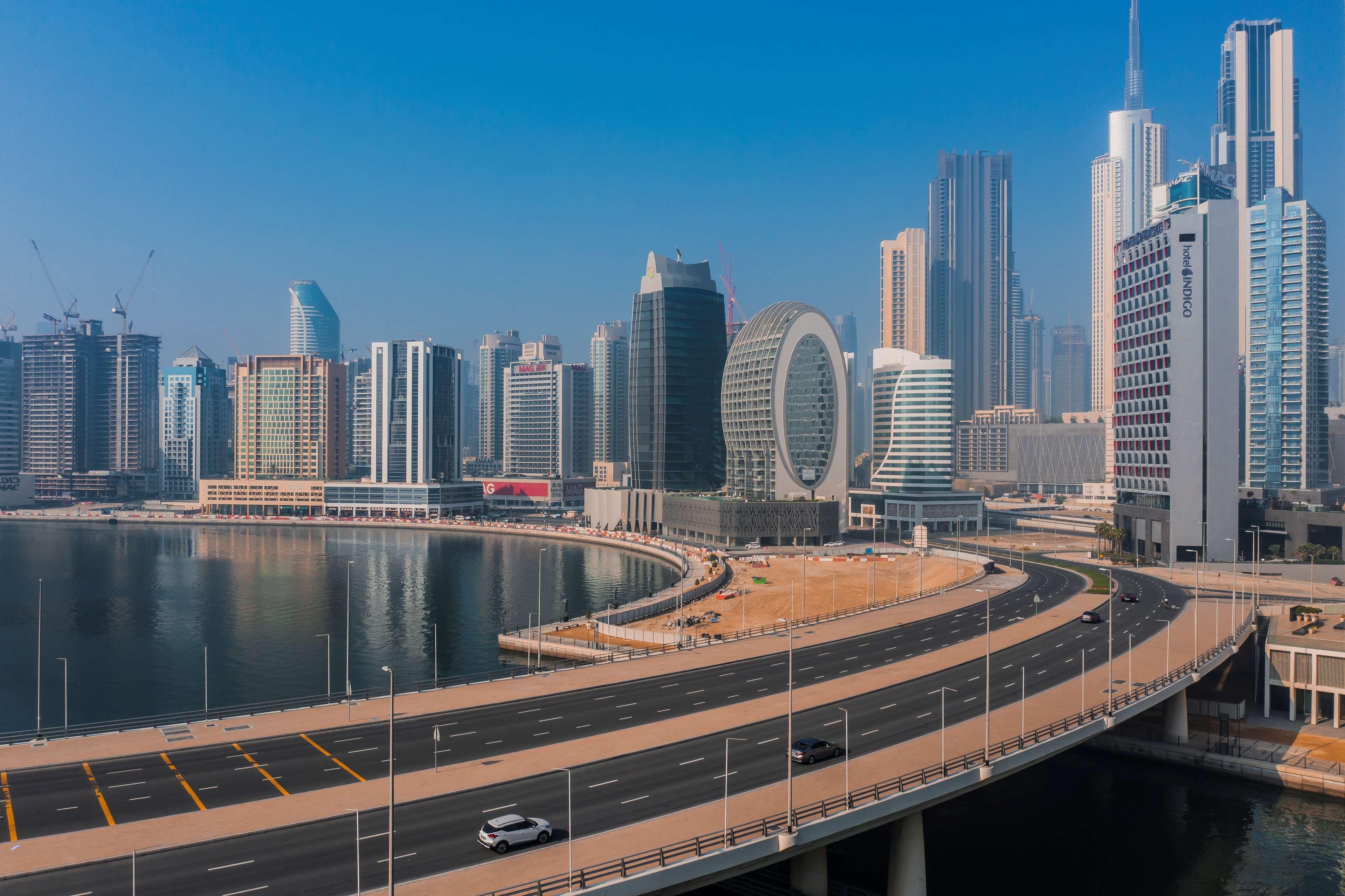 Radisson Blu Hotel, Dubai Canal View Exterior photo The image depicts a modern city skyline featuring a variety of tall buildings. In the foreground, there is a curved roadway that runs alongside a body of water, reflecting the surrounding architecture. The skyline is characterized by a mix of contemp