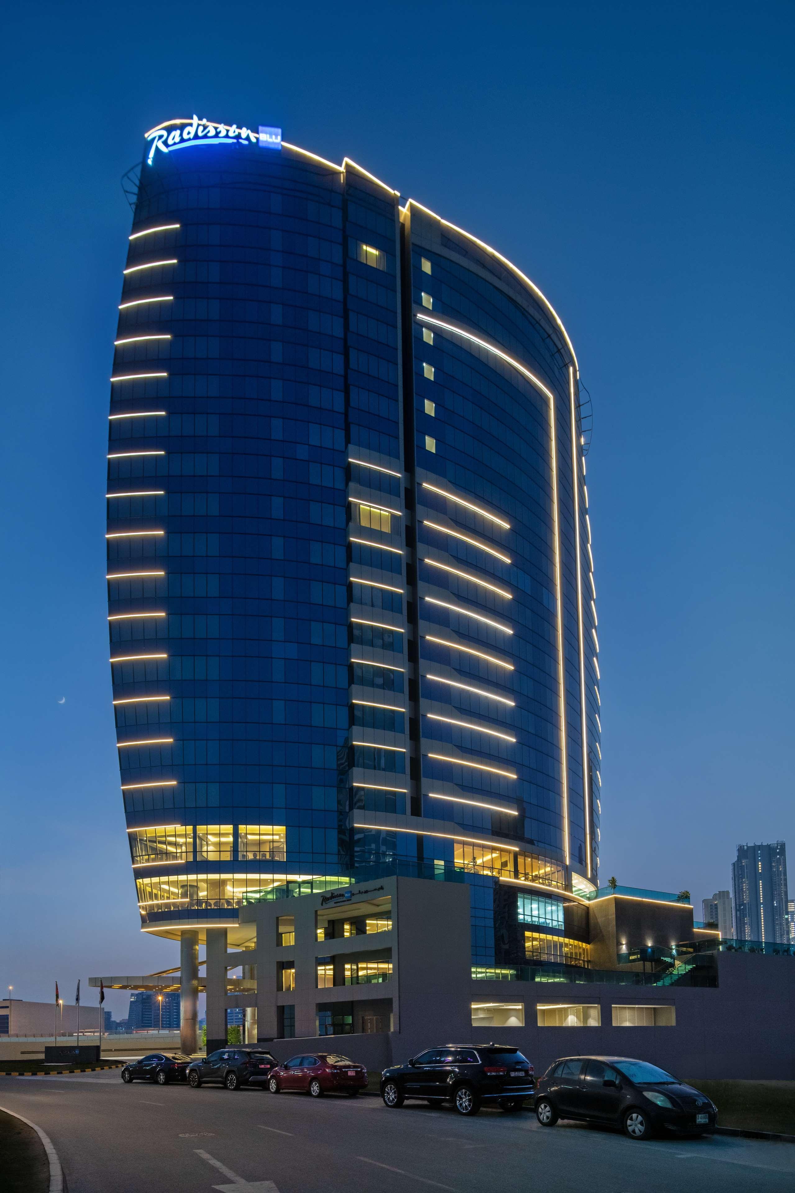 Radisson Blu Hotel, Dubai Canal View Exterior photo The image shows a modern skyscraper with a sleek, curved design. The building is primarily covered in glass, reflecting the evening sky, and features illuminated lines along its surface, enhancing its architectural appeal. The lighting creates a stri