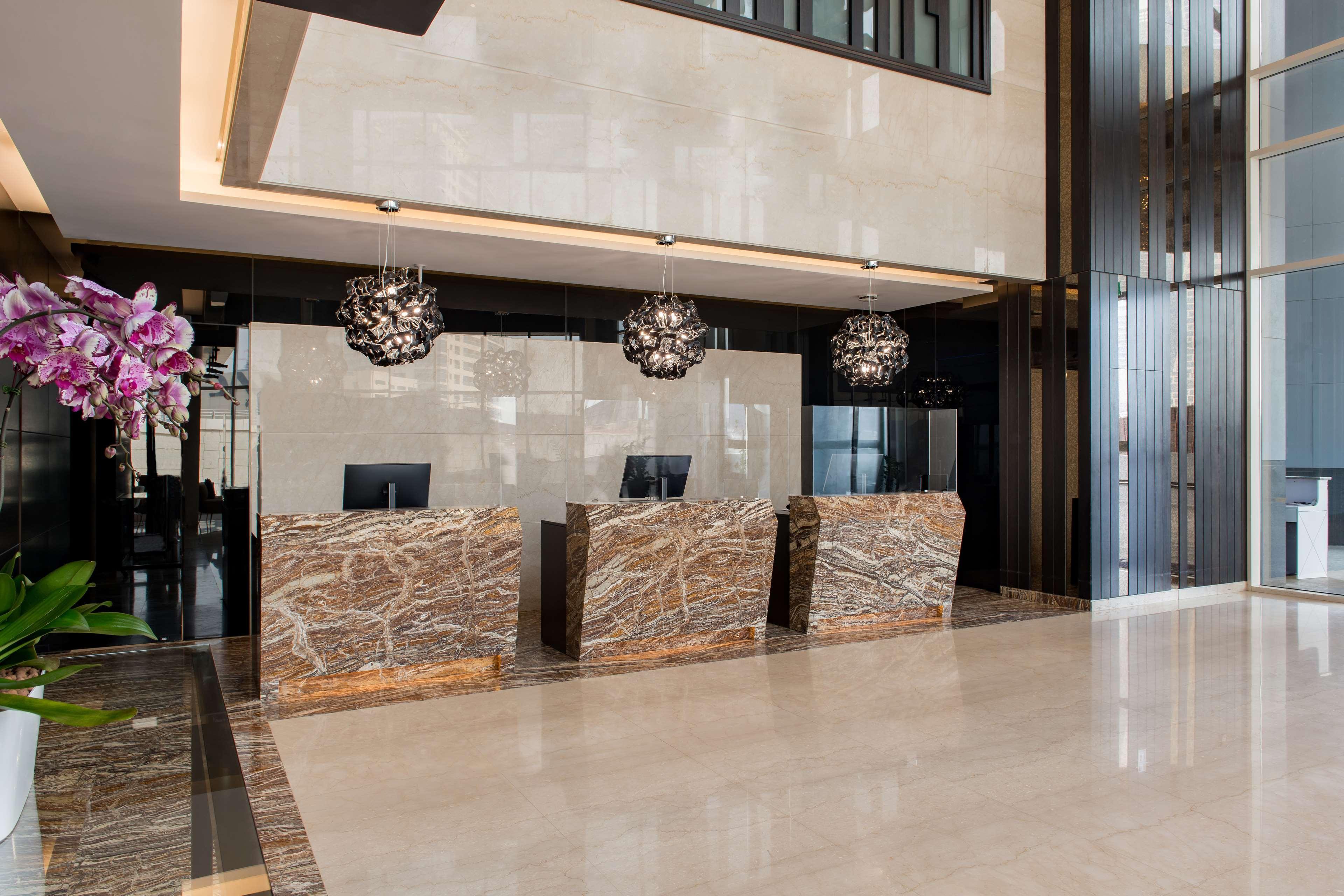 Radisson Blu Hotel, Dubai Canal View Exterior photo The photo shows a modern reception area. There are three reception counters made of elegant marble, facing the viewer, with a sleek and minimalist design. Above each counter, there are decorative hanging light fixtures that add a contemporary touch. 