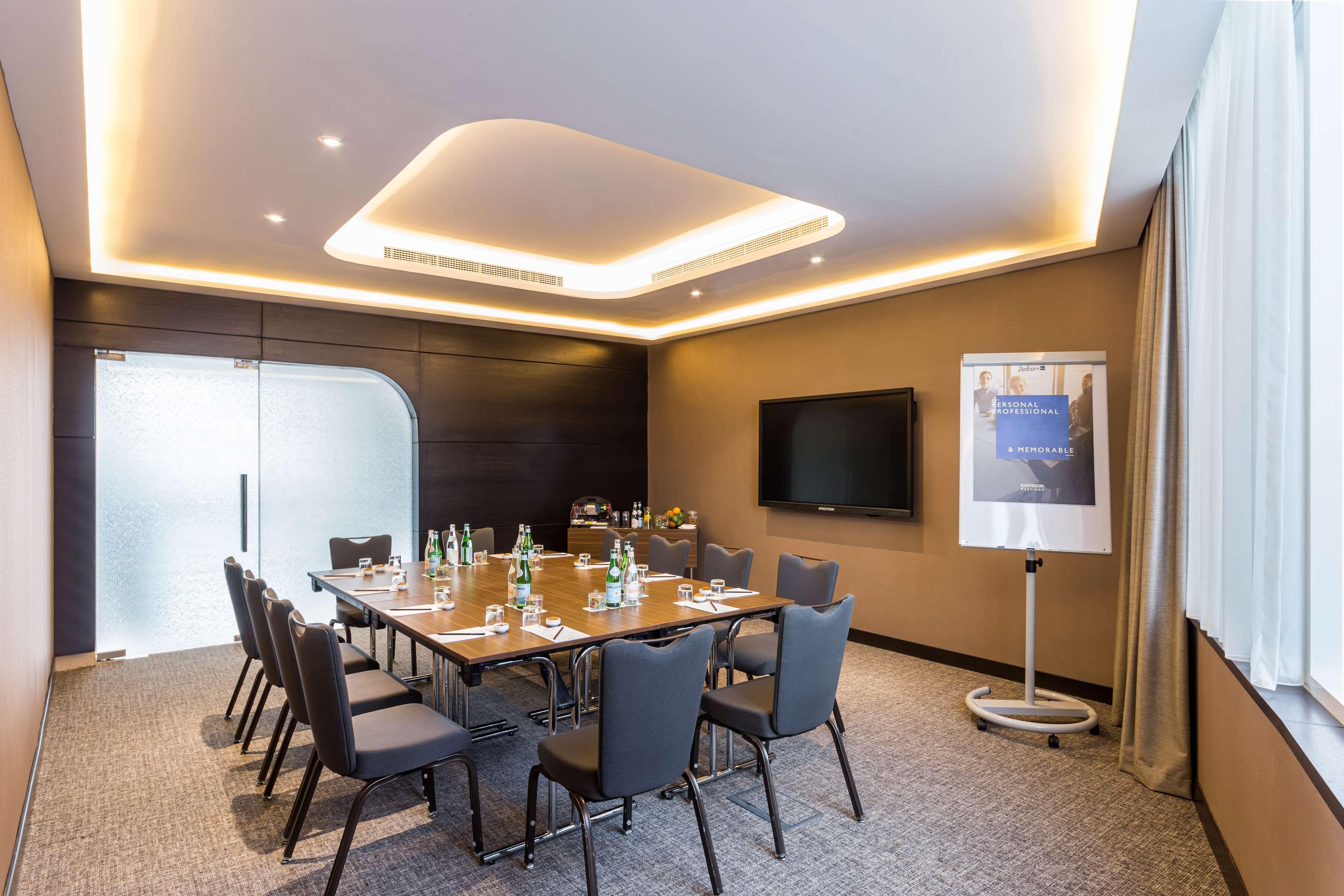 Radisson Blu Hotel, Dubai Canal View Exterior photo The photo shows a modern conference room. It features a large wooden table with several chairs arranged around it. On the table, there are multiple bottles of water, indicating it's set up for a meeting. The room has a large wall-mounted television a