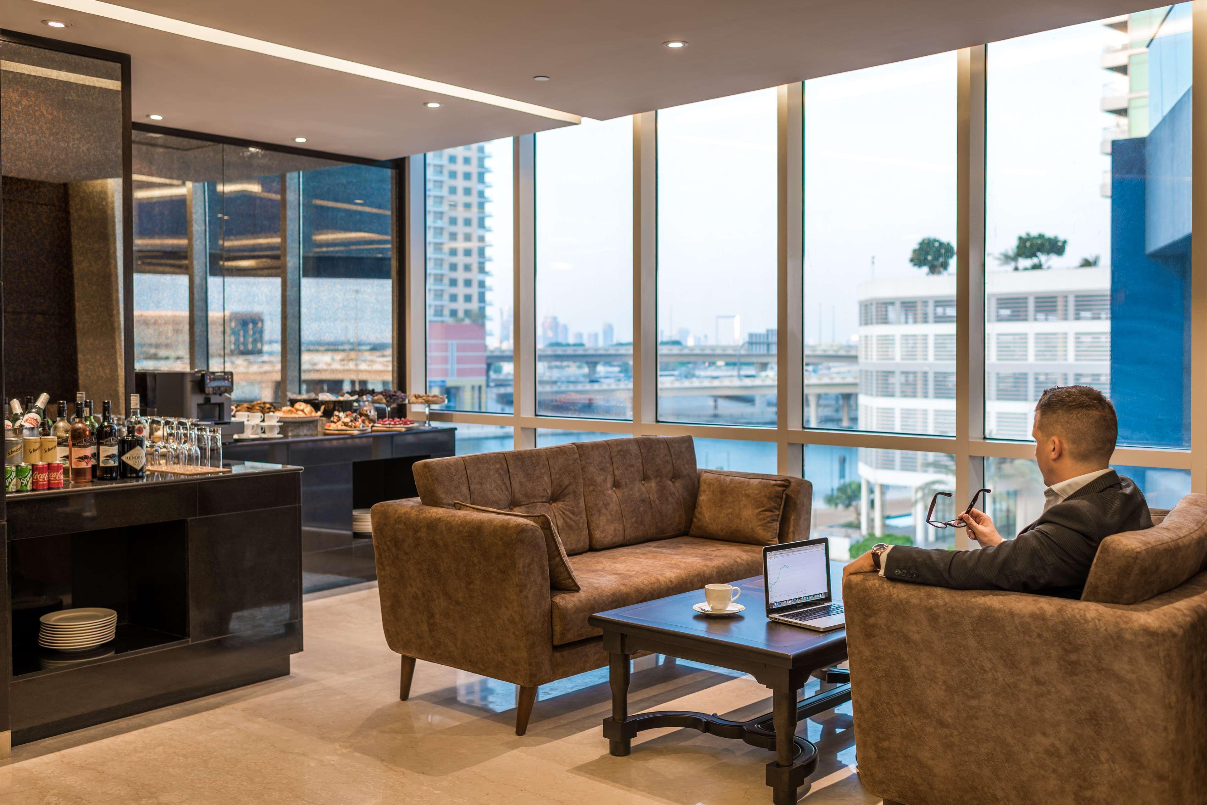 Radisson Blu Hotel, Dubai Canal View Exterior photo The photo depicts a modern and stylish lounge area with large windows that offer a view of the cityscape. There are two brown upholstered sofas and a wooden table situated in the center. On the table, there is a laptop, a coffee cup, and a smartphone