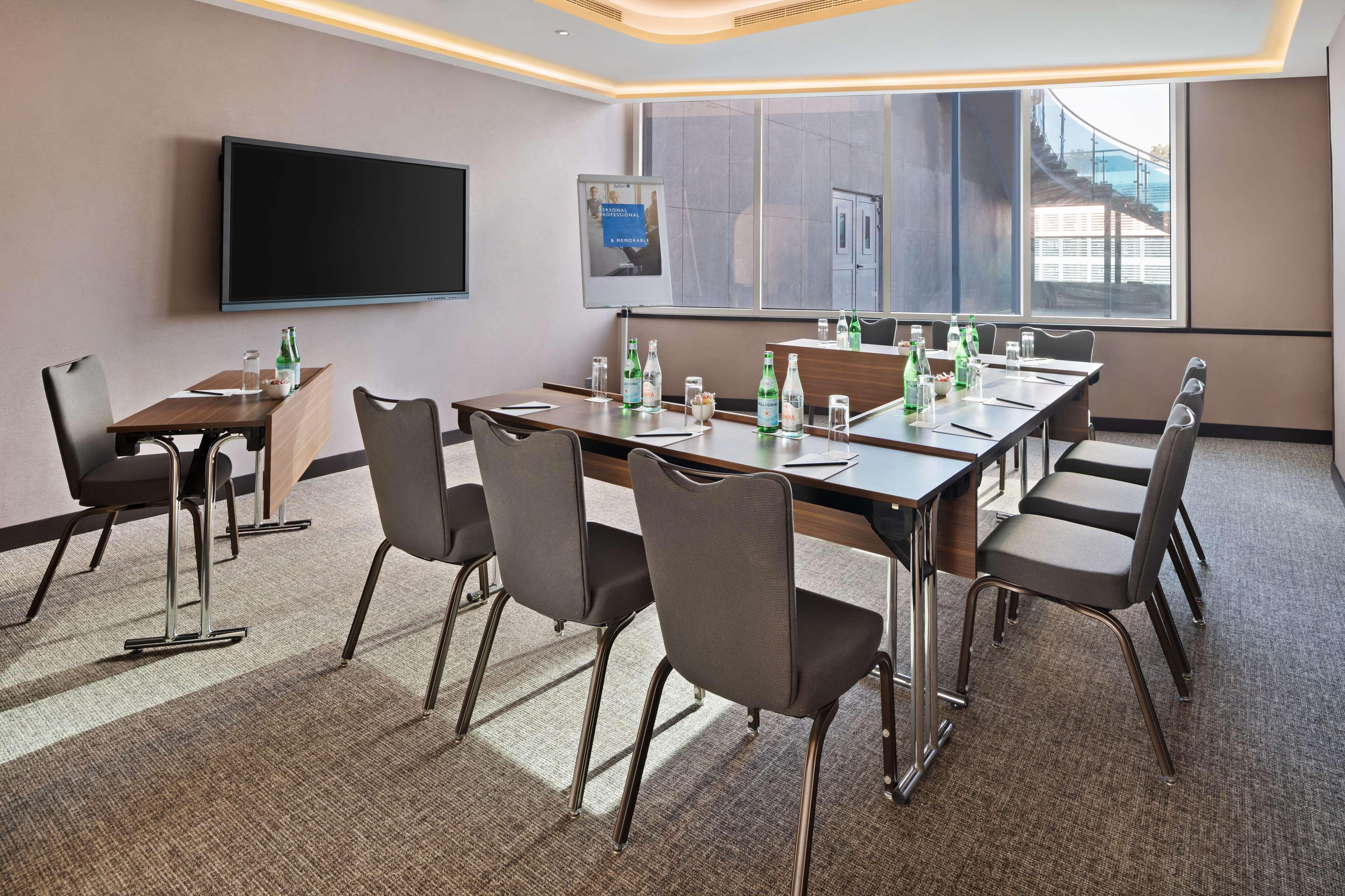 Radisson Blu Hotel, Dubai Canal View Exterior photo The photo shows a modern conference room equipped for meetings. There is a large rectangular table surrounded by several chairs. On the table, there are several glass water bottles and glasses placed for participants. A large flat-screen TV is mounte