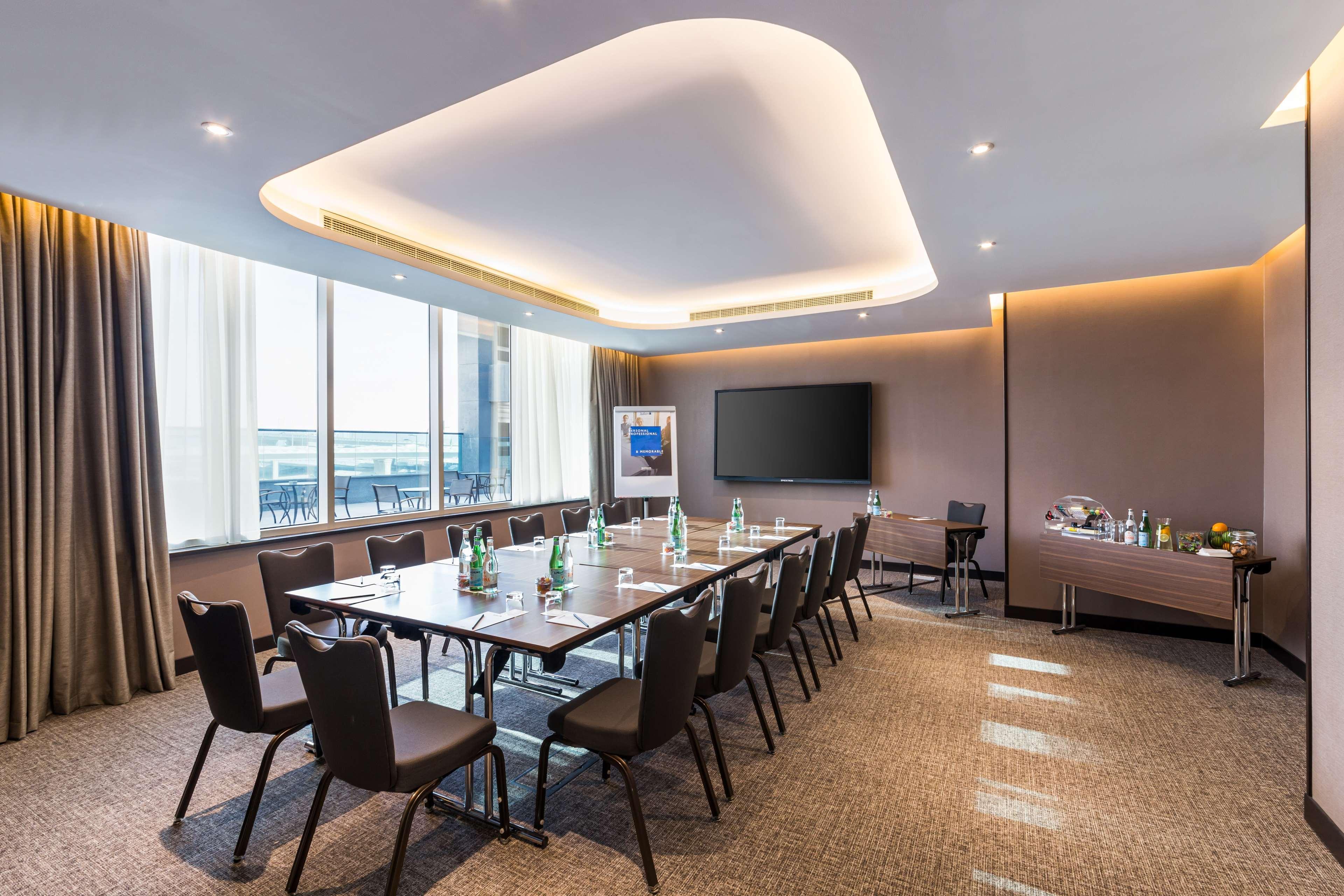 Radisson Blu Hotel, Dubai Canal View Exterior photo The photo shows a modern conference room. It features a long rectangular table surrounded by black chairs. There are several bottles of water on the table, indicating a setup for a meeting. The room has large windows that allow natural light to enter