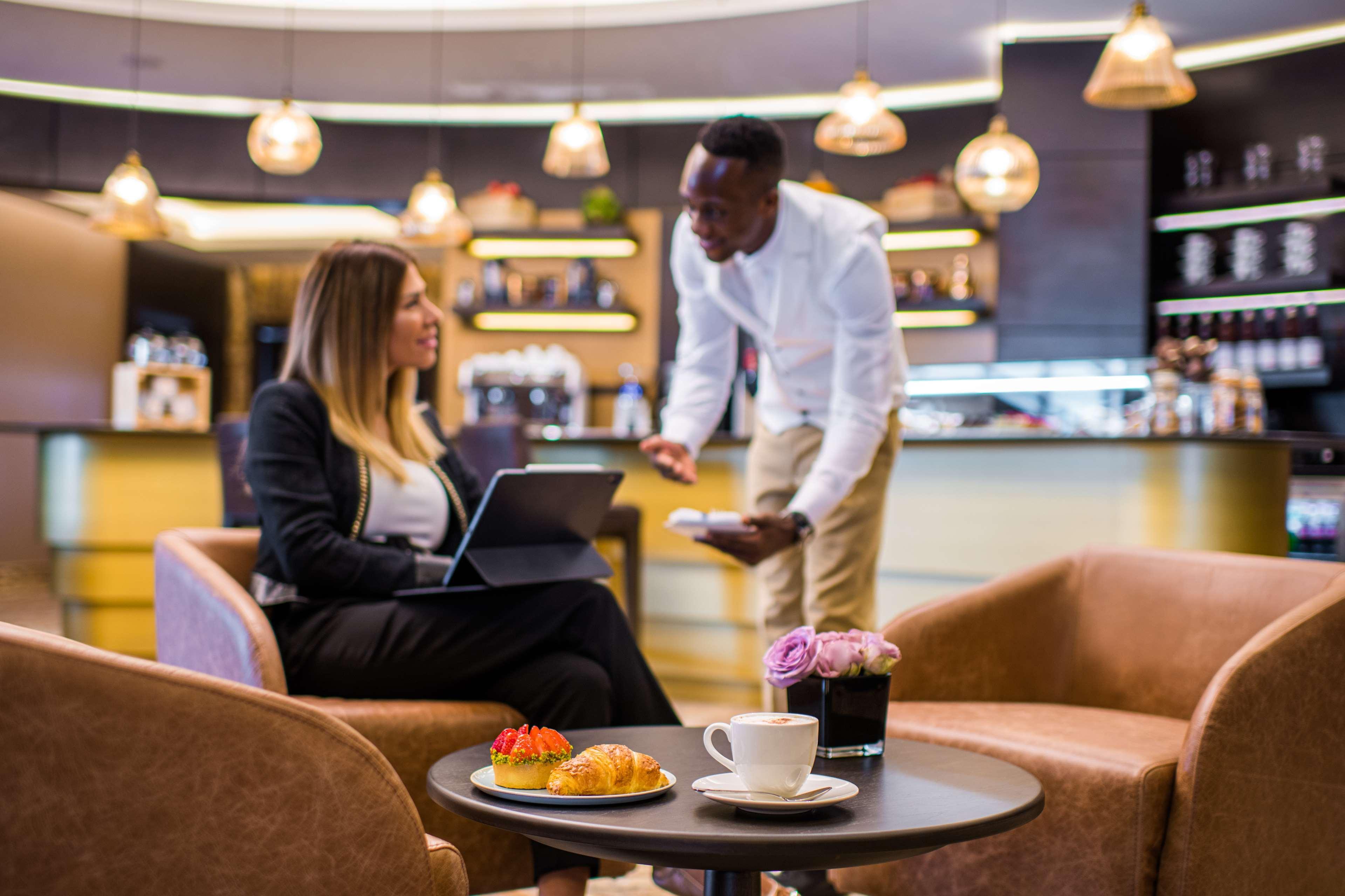 Radisson Blu Hotel, Dubai Canal View Exterior photo The photo depicts a cozy café or lounge setting. In the foreground, a woman is seated in an armchair, working on a laptop and engaging with a male server who is standing beside her. The server is holding a tray, likely offering her food or drink. The