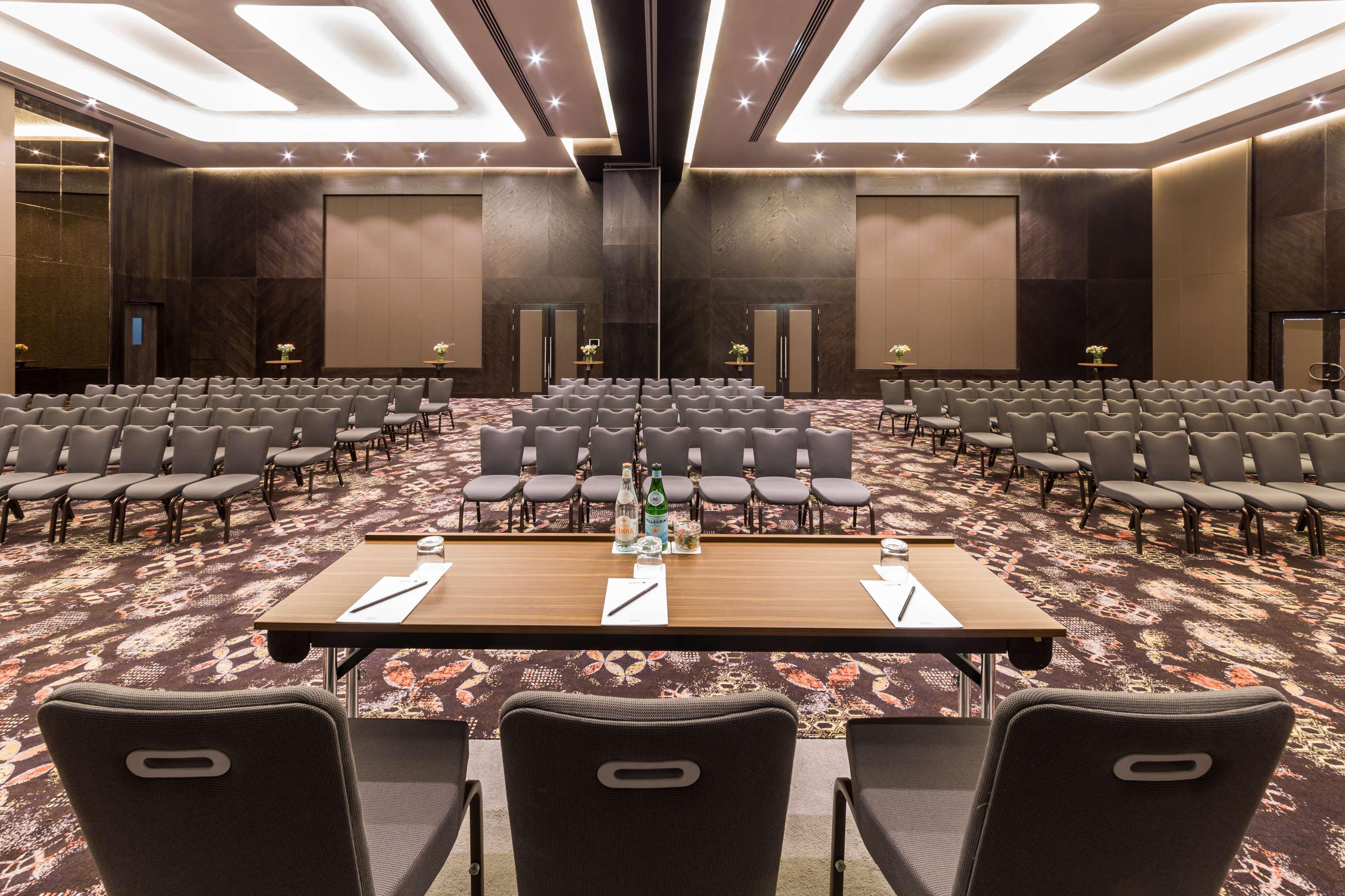 Radisson Blu Hotel, Dubai Canal View Exterior photo The photo depicts an elegant conference or meeting room. In the foreground, there is a wooden table with some water bottles and papers, likely for speakers or panelists. Directly behind the table, there are rows of gray chairs arranged neatly, indica