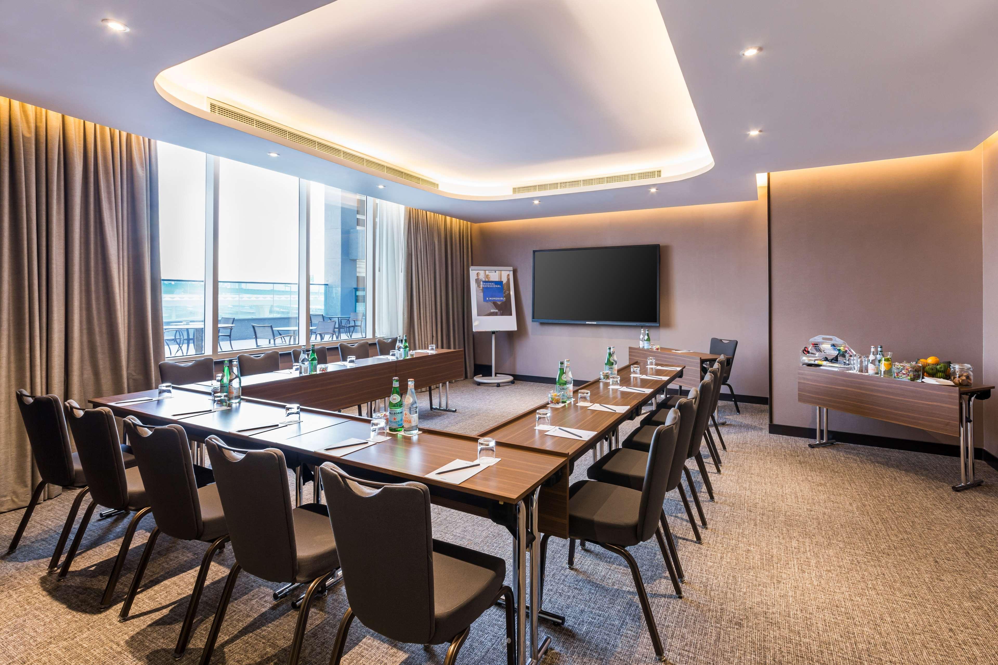 Radisson Blu Hotel, Dubai Canal View Exterior photo The photo shows a modern conference room arranged for a meeting. There are several tables set up in a U-shape, with comfortable black chairs around them. On each table, there are glass bottles of water, small glasses, and some note-taking materials. 