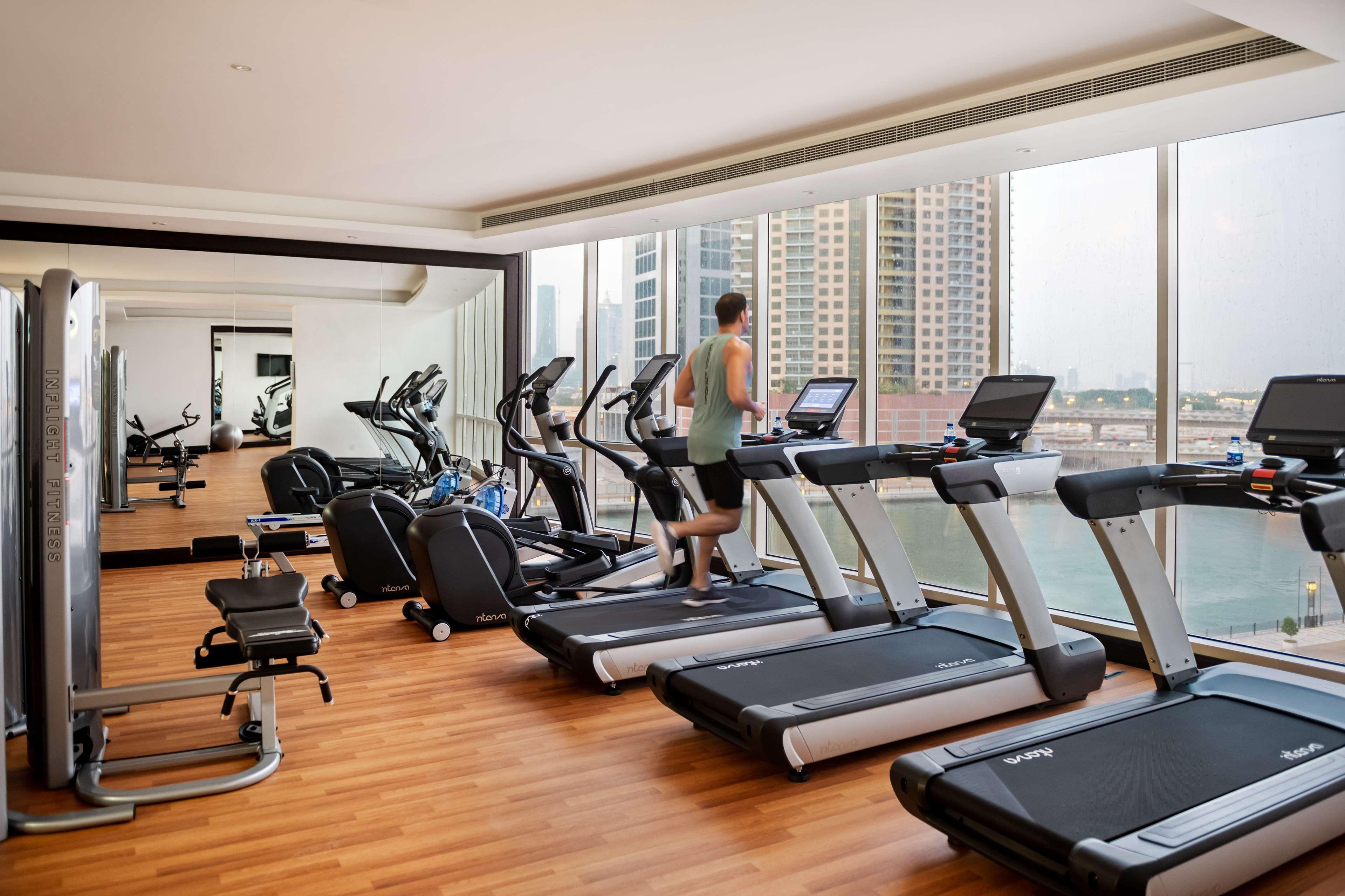 Radisson Blu Hotel, Dubai Canal View Exterior photo The photo shows a modern gym with plenty of natural light. There are several exercise machines, including treadmills and elliptical machines, arranged along one side of the room. A person is running on one of the treadmills. The gym features a wooden