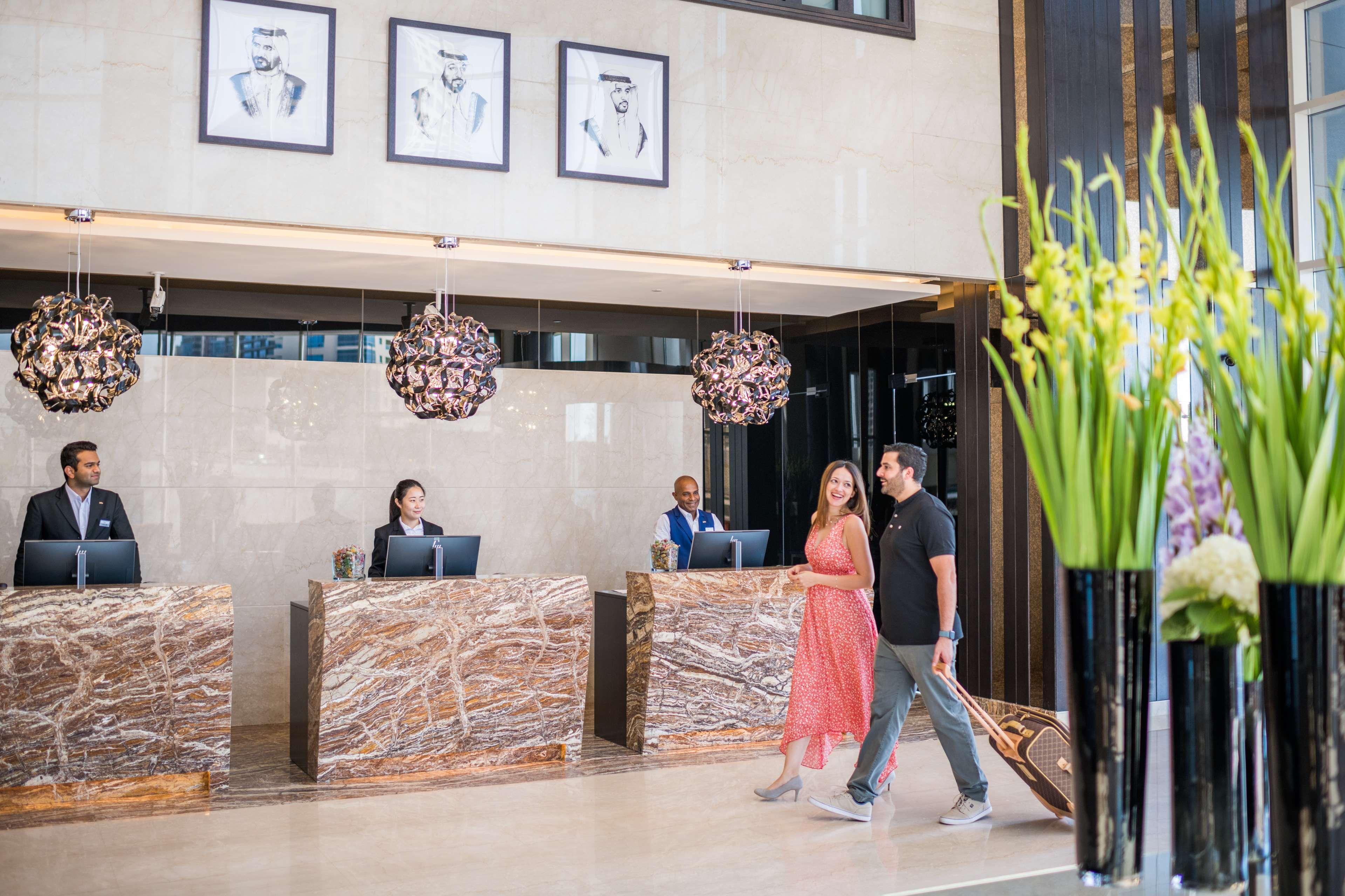 Radisson Blu Hotel, Dubai Canal View Exterior photo The photo shows a stylish hotel lobby or reception area. In the foreground, a couple is walking past the reception desk, with the woman wearing a pink dress. The reception area features elegant marble counters, and behind them, staff members are atte