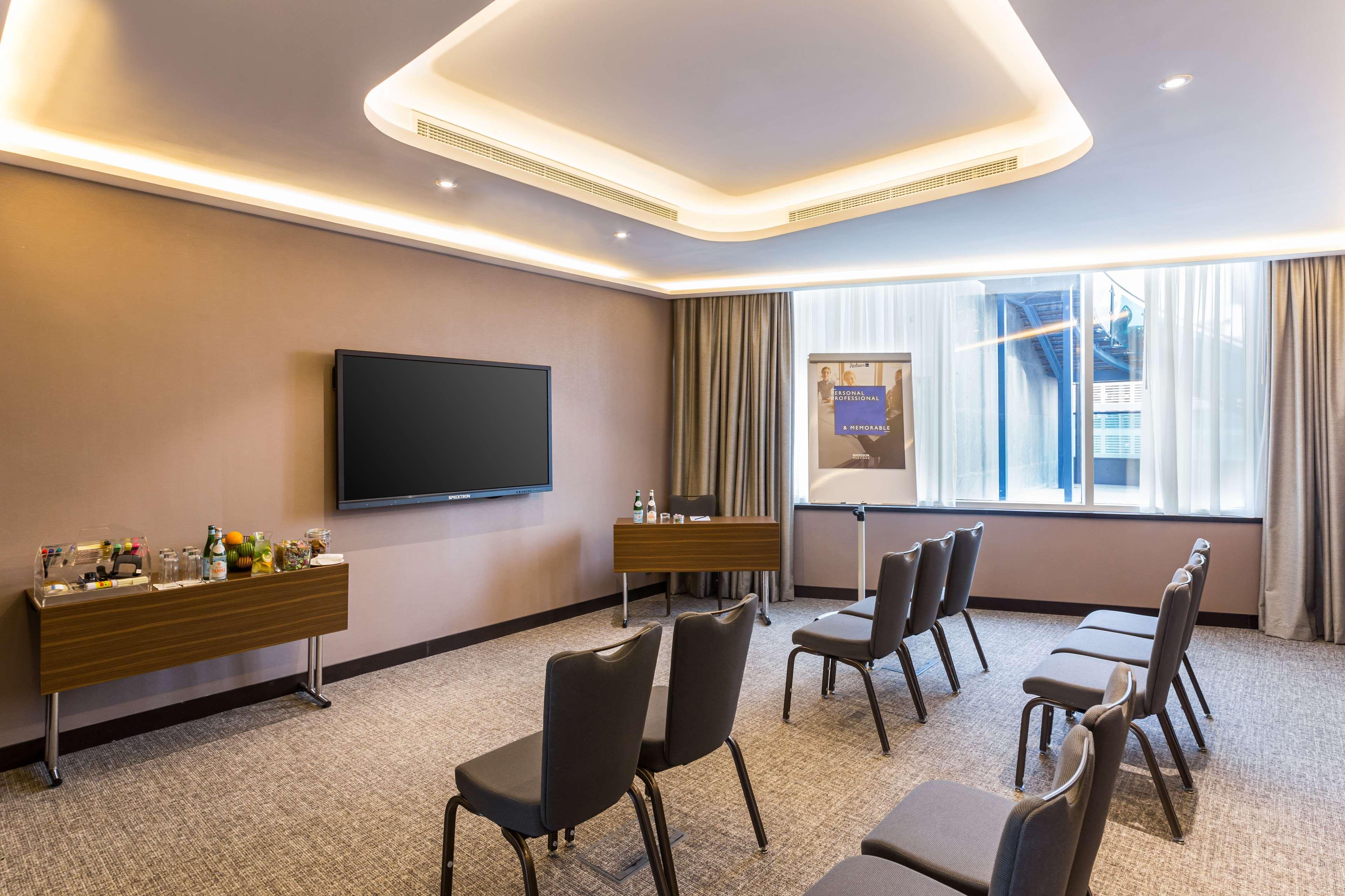 Radisson Blu Hotel, Dubai Canal View Exterior photo The photo shows a modern meeting room. It features several rows of chairs arranged facing a large wall-mounted television. There's a wooden table or console on one side that holds some refreshments. The room has soft lighting and large windows with c