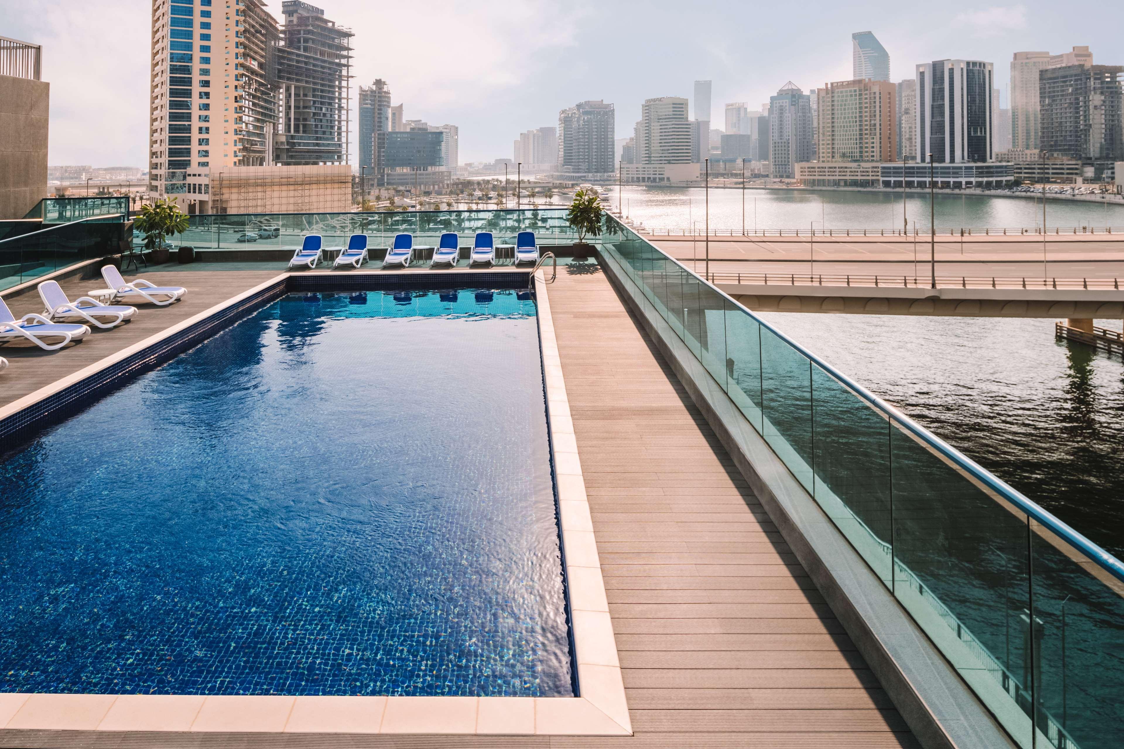 Radisson Blu Hotel, Dubai Canal View Exterior photo The photo shows a scenic view of a pool area overlooking a city skyline. The swimming pool is rectangular and features a shimmering blue surface, surrounded by lounge chairs for relaxation. There are glass railings providing a clear view of the surro