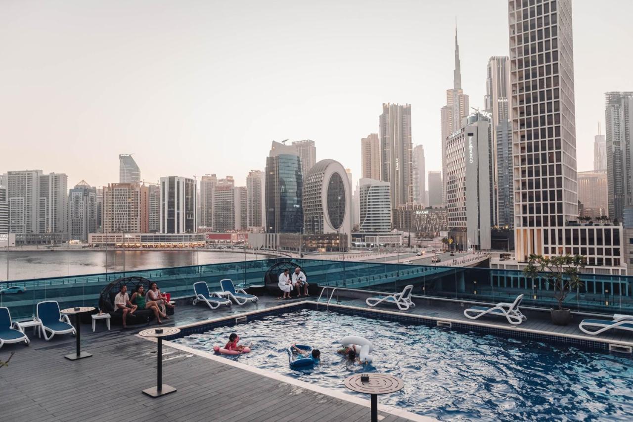 Radisson Blu Hotel, Dubai Canal View Exterior photo The photo shows a rooftop pool area with several lounge chairs arranged around it. In the pool, there are a few people enjoying themselves. In the background, a city skyline is visible, featuring tall modern buildings, including a prominent skyscrape