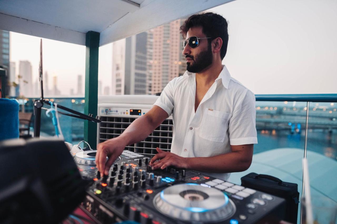 Radisson Blu Hotel, Dubai Canal View Exterior photo The photo shows a DJ playing music at an outdoor venue, likely on a rooftop or terrace. He is wearing sunglasses and a casual white shirt. The DJ is focused on his equipment, which includes a DJ mixer and turntables. In the background, there are tall