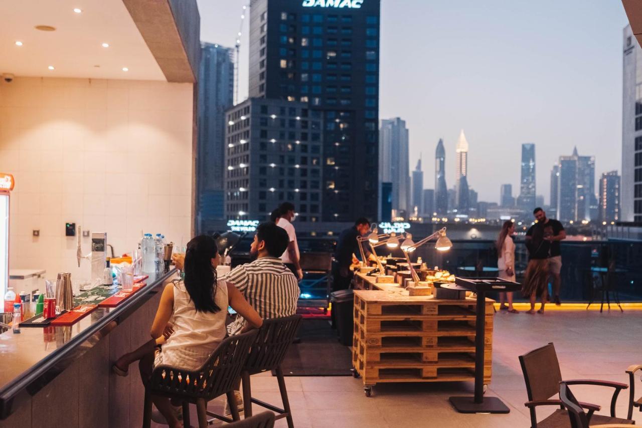 Radisson Blu Hotel, Dubai Canal View Exterior photo The photo depicts a rooftop setting during twilight. In the foreground, a couple is seated at the bar area, while in the background, a buffet table made of wooden pallets showcases various food items. Several people can be seen mingling, with a city 